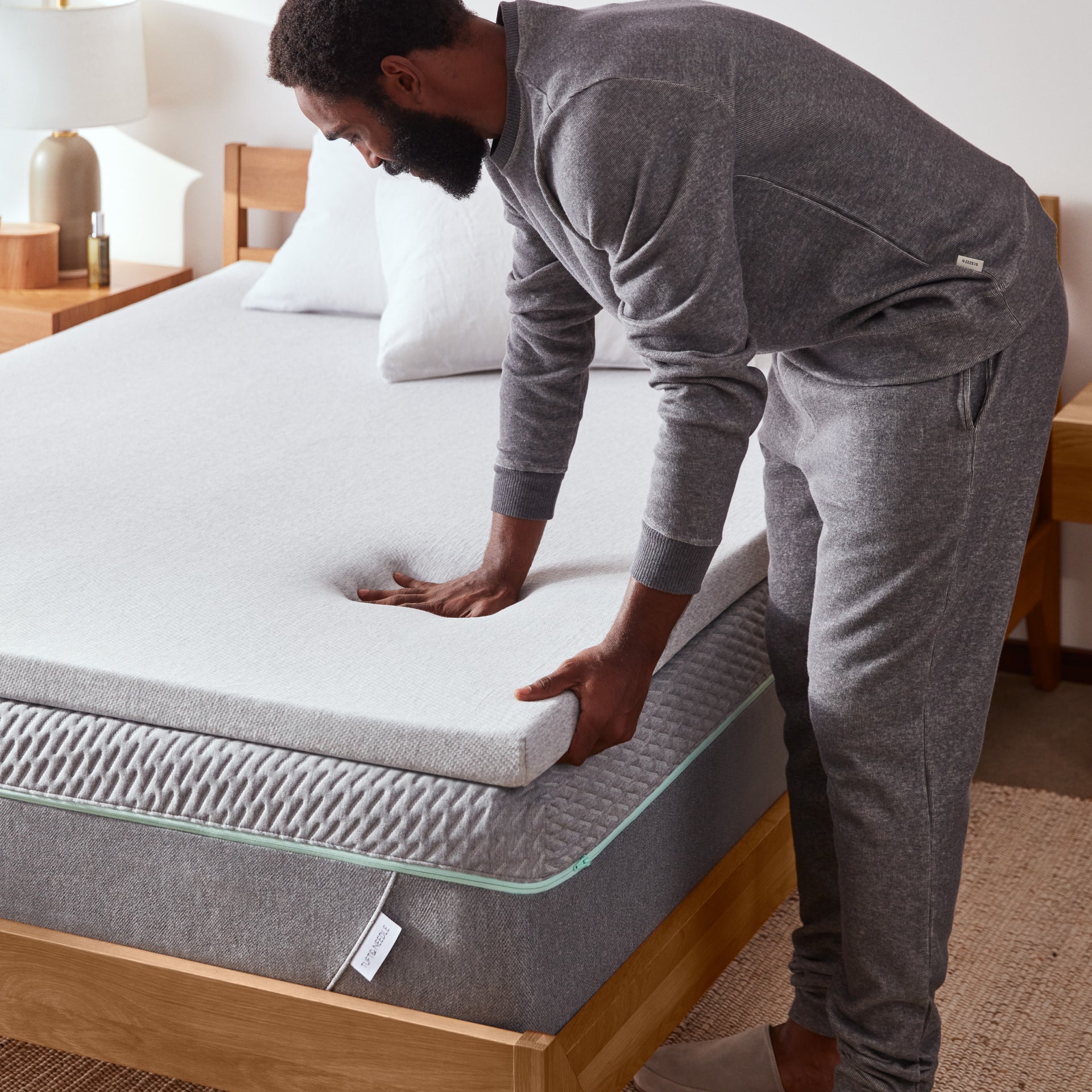 A man presses down on a soft, grey mattress topper layered over a textured mattress, testing its comfort and responsiveness.